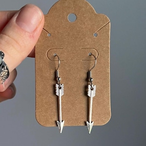 May include: A pair of silver arrow earrings on a brown card. The earrings are simple and elegant, with a small arrow charm hanging from a silver hook.