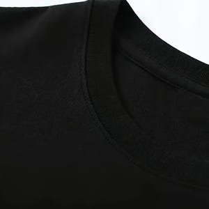 Puede incluir: Primer plano de una camiseta negra con cuello redondo. La tela parece suave y lisa, con un cuello y una costura del hombro visibles. La imagen est&aacute; bien iluminada, destacando la textura y el color de la prenda.