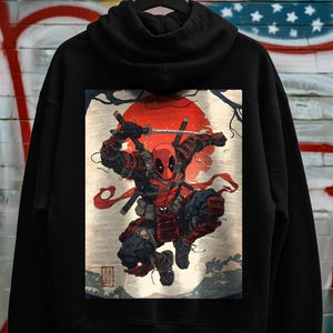 May include: Black hoodie featuring a vibrant graphic of Deadpool in a samurai-inspired pose, set against a red sun and traditional Japanese art elements. The design includes swords and a dynamic action stance.