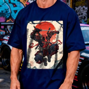 Puede incluir: Camiseta azul marino con un dise&ntilde;o de Deadpool en una pose inspirada en los samur&aacute;is, con un sol rojo y texto japon&eacute;s. El dise&ntilde;o incluye espadas y un disfraz detallado. Una prenda de vestir informal.
