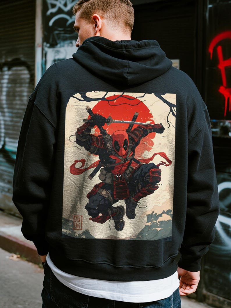 May include: Black hoodie featuring a graphic of Deadpool in a samurai-inspired design. The artwork includes a red sun, swords, and Japanese-style elements. The hoodie is made of a soft material and has a hood and a front pocket.