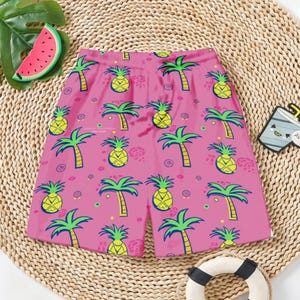 May include: Pink shorts with a repeating pattern of yellow pineapples and green palm trees. The shorts have an elastic waistband and are likely made of a lightweight fabric, perfect for warm weather.