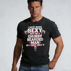Puede incluir: Camiseta negra con el texto "I HATE BEING SEXY BUT I'M A CHUBBY BEARDED MAN SO I CAN'T HELP IT" en blanco y rojo. El diseño incluye hachas y una calavera. La camiseta la lleva una persona.