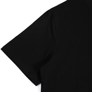 Puede incluir: Una camiseta negra lisa de manga corta. La imagen muestra el hombro y la manga de la camiseta sobre un fondo blanco. La camiseta est&aacute; hecha de una tela lisa y opaca. El dise&ntilde;o es simple y cl&aacute;sico.