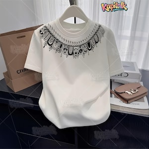 May include: A white short-sleeved t-shirt featuring a black graphic print around the neckline. The design consists of a repeating pattern of stylized arches and intricate line work. The shirt is displayed on a hanger and includes the text Kapitalk.