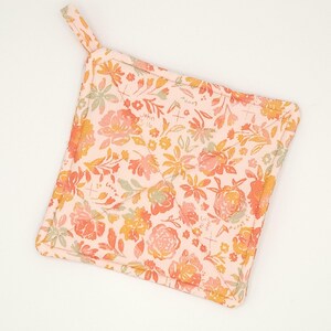 May include: A square, floral-patterned pot holder in shades of peach, orange, yellow, and green. The fabric features a repeating pattern of flowers and leaves, with the text "John 3:16" visible. A small loop is attached for hanging.