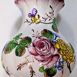 Puede incluir: Un jarrón de cerámica blanca con un borde con volantes, adornado con diseños florales pintados a mano. Las flores incluyen una gran rosa rosa, flores amarillas y azules y hojas verdes. El borde es de color burdeos oscuro.