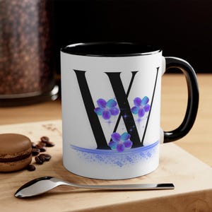 May include: White ceramic mug with a black interior and handle. The letter "W" is displayed in black, decorated with purple and blue floral accents. A blue decorative line and sparkles complete the design.