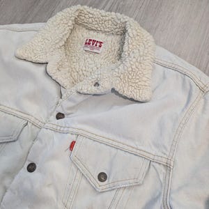 Könnte beinhalten: Hellblaue Jeansjacke mit cremefarbenem Sherpa-Kragen. Die Jacke hat zwei Brusttaschen, Knopfverschlüsse und das Levi's-Markenetikett. Ein klassisches, lässiges Oberbekleidungsstück.