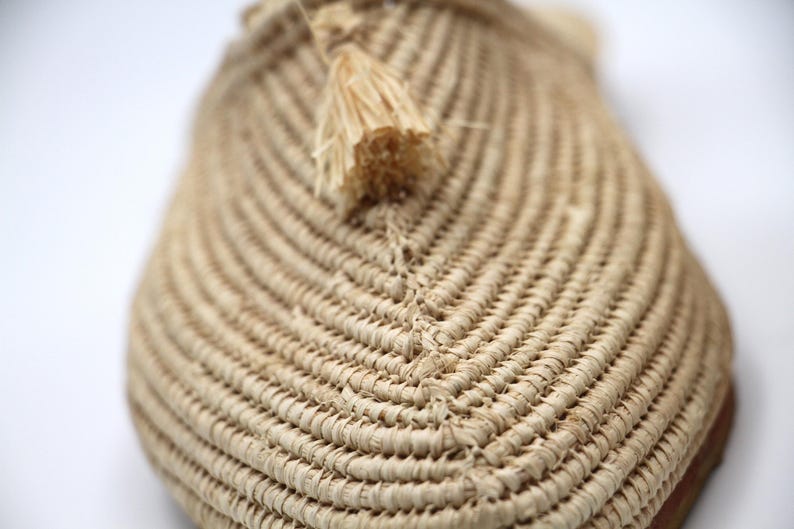 K&ouml;nnte beinhalten: Nahaufnahme eines geflochtenen Strohslippes mit einem kleinen Quasten. Der Slipper ist beige und hat ein nat&uuml;rliches, rustikales Aussehen.