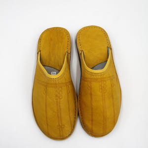 Puede incluir: Un par de zapatillas de cuero amarillo mostaza. Las zapatillas tienen una puntera redondeada y un diseño sin cordones. Líneas verticales decorativas y motivos florales están grabados en la parte superior de cada zapatilla. Los bordes están cosidos con hilo en contraste.