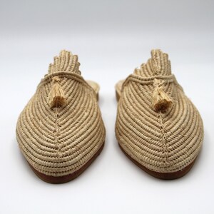 K&ouml;nnte beinhalten: Ein Paar beige geflochtene Strohslipper mit Quasten. Die Slipper haben eine spitze Zehenpartie und eine flache Sohle.