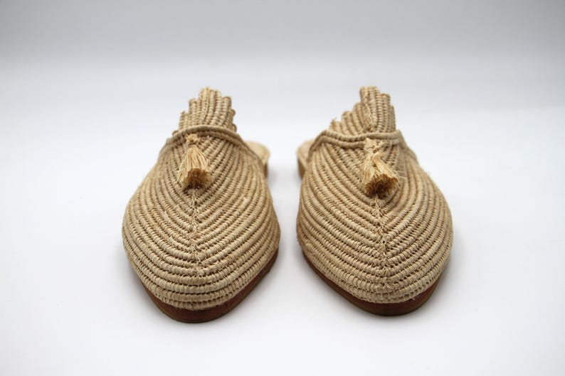 K&ouml;nnte beinhalten: Ein Paar beige geflochtene Strohslipper mit Quasten. Die Slipper haben eine spitze Zehenpartie und eine flache Sohle.