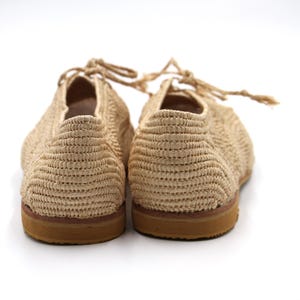 May include: A pair of beige woven espadrille shoes with brown soles. The shoes have a lace-up design and are made from a natural material.
