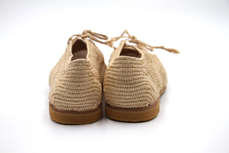 May include: A pair of beige woven espadrille shoes with brown soles. The shoes have a lace-up design and are made from a natural material.