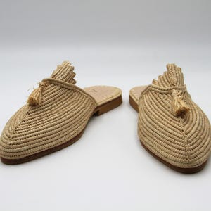 K&ouml;nnte beinhalten: Ein Paar beige geflochtene Strohslipper mit Quasten. Die Slipper haben eine spitze Zehenpartie und eine flache Sohle.