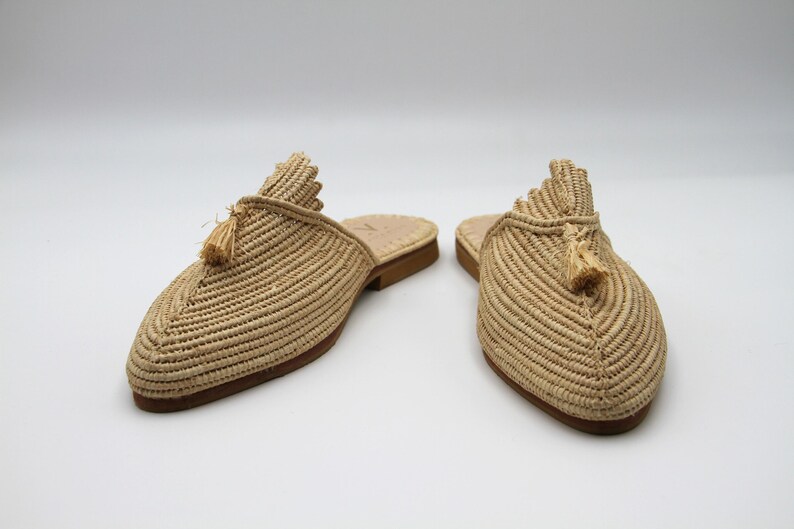 K&ouml;nnte beinhalten: Ein Paar beige geflochtene Strohslipper mit Quasten. Die Slipper haben eine spitze Zehenpartie und eine flache Sohle.