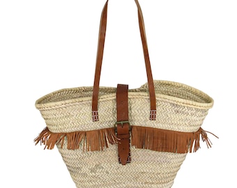 Puede incluir: Un bolso tote de paja tejido con una correa de cuero marrón y ribete de flecos. El bolso tiene un cierre de hebilla y es perfecto para llevar los artículos esenciales del día a día.