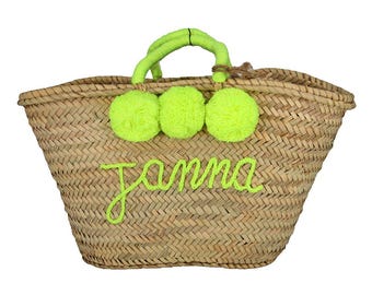 May include: A straw tote bag with a bright green handle and pom poms. The bag is personalized with the name "Jamma" in green thread.