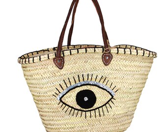 Puede incluir: Un gran bolso de paja con asas de cuero marrón y un diseño de ojo malvado en blanco y negro con detalles de purpurina plateada.