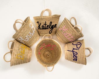 May include: Woven straw tote bags with embroidered names in various colors. The bags have small handles and are arranged on a white surface. The names include Katelyn, Mattie, Piper, and others.
