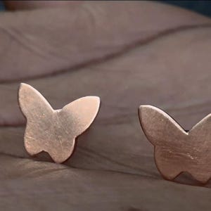May include: Two copper-colored butterfly stud earrings. The earrings are small, with a simple butterfly silhouette. The metal has a slightly textured surface. The earrings are displayed on a neutral background.