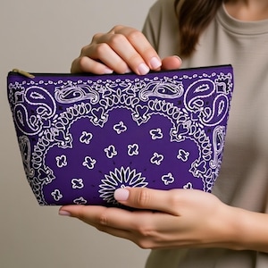 Könnte beinhalten: Eine lila Kosmetiktasche mit weißem Paisley-Muster. Die Tasche hat einen schwarzen Reißverschluss und einen goldfarbenen Reißverschlussanhänger. Die Tasche wird von einer Person gehalten.