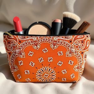 May include: Orange cosmetic bag with a paisley pattern, containing makeup items like lipstick, powder, and brushes. The bag has a black zipper and is filled with various beauty products.