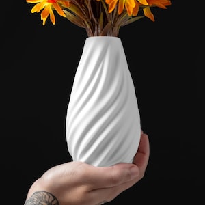 May include: A white, spiral-textured vase holds orange and yellow flowers. The vase is held in a hand against a black background. The vase has a unique, swirling design, and the flowers add a pop of color.