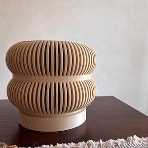 May include: A beige, ribbed vase with a rounded shape. The vase has a cylindrical base and two stacked, bulbous sections, each with vertical grooves. The design is modern and minimalist, suitable for home decor.