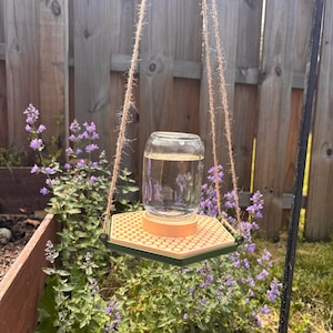 May include: A hanging bird feeder with a clear glass jar filled with water. The feeder has a hexagonal tray with a textured surface and a green border. The feeder is suspended by twine ropes, with purple flowers and a wooden fence in the background.