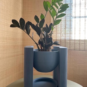 May include: A ZZ plant with dark green and black leaves potted in a matte blue bowl-shaped planter. The planter is suspended within a blue vertical stand featuring a ribbed texture on the exterior columns and a circular base tray underneath.