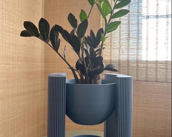 May include: A ZZ plant with dark green and black leaves potted in a matte blue bowl-shaped planter. The planter is suspended within a blue vertical stand featuring a ribbed texture on the exterior columns and a circular base tray underneath.