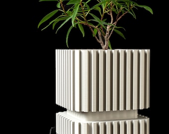 May include: A white square planter with a vertical ribbed texture featuring a recessed middle section. The planter has a two-tiered design with a wider top section and a narrower base. A small green leafy plant grows out of the top of the planter.