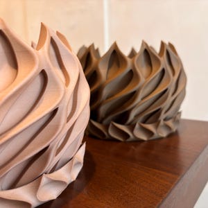 May include: Two decorative, textured vases. One is a light brown color, and the other is a darker brown. The vases have a unique, wave-like design with pointed edges. They are placed on a dark wooden surface.