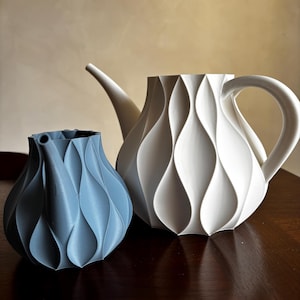 May include: Two decorative vases with a unique, textured design. One is a light blue color, and the other is white. The vases have a wavy, vertical pattern. The white vase has a spout and handle, resembling a teapot. The vases are on a dark wooden surface.