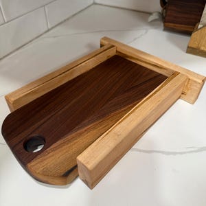Pode incluir: Uma tábua de corte de madeira com um centro castanho escuro e uma moldura castanha clara. A tábua tem uma extremidade arredondada com um orifício e foi concebida para deslizar para dentro de uma moldura de madeira. A tábua de corte é feita de dois tipos diferentes de madeira.