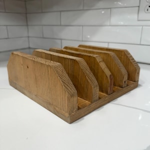 May include: A wooden desk organizer with five vertical slots. The organizer is made of light brown wood and has a rectangular base. The slots are angled and provide storage for mail, documents, or other office supplies.