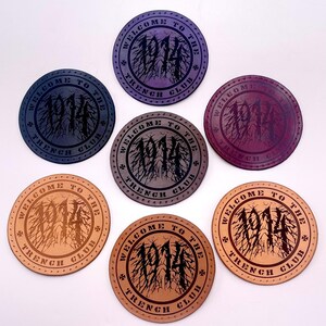 Peut inclure: Sept dessous de verre circulaires avec l'inscription "WELCOME TO THE TRENCH CLUB" et l'année "1914". Les dessous de verre sont de couleurs variées, notamment violet, marron et gris, avec un motif de racines d'arbre.