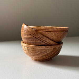 May include: Three small, wooden bowls stacked on top of each other. The bowls are made of light brown wood with visible grain and a smooth finish. Each bowl has a rounded shape with a slightly flared rim, perfect for serving small portions.