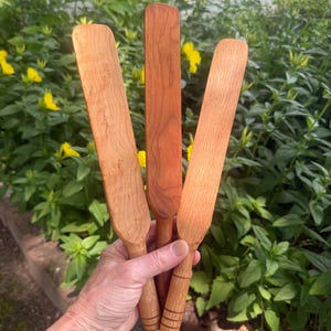 May include: Three wooden kitchen spatulas with long, flat blades and rounded handles. The spatulas feature distinct wood grain patterns in varying shades of light to medium brown, with one handle showing three horizontal engraved lines.