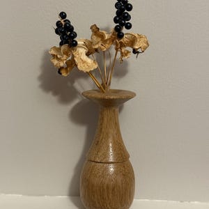 May include: A small, wooden vase with a bulbous base and a narrow neck, holding dried seed pods and clusters of black berries. The vase is a light brown color and sits on a white shelf against a neutral wall.