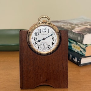 May include: A vintage Hamilton pocket watch with a white face and gold-tone casing, displayed in a dark wooden stand. The watch features black numerals and hands, with a smaller seconds dial. The stand has a curved base and a cutout to hold the watch.
