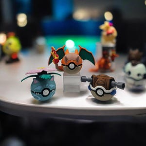 May include: Three spherical character figurines featuring a blue creature with a pink flower, an orange creature with wings and a flame, and a brown creature with two cannons. Each figure is integrated into a white and black circular ball design.