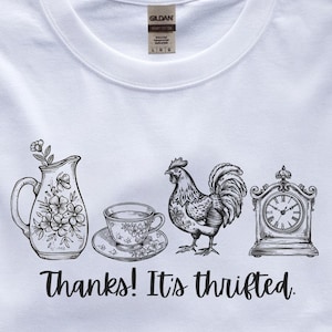 May include: White t-shirt with a black graphic design featuring a floral pitcher, teacup, rooster, and clock. The text "Thanks! It's thrifted" is printed below the illustrations. The shirt is made of a soft, comfortable material.