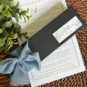 Puede incluir: Una caja negra con una cinta azul claro y una tarjeta con texto. La caja tiene una etiqueta blanca con dise&ntilde;os florales y las palabras "La Belle Handmade". La tarjeta dice "Blessing Sash: Mother/Daughter Edition".