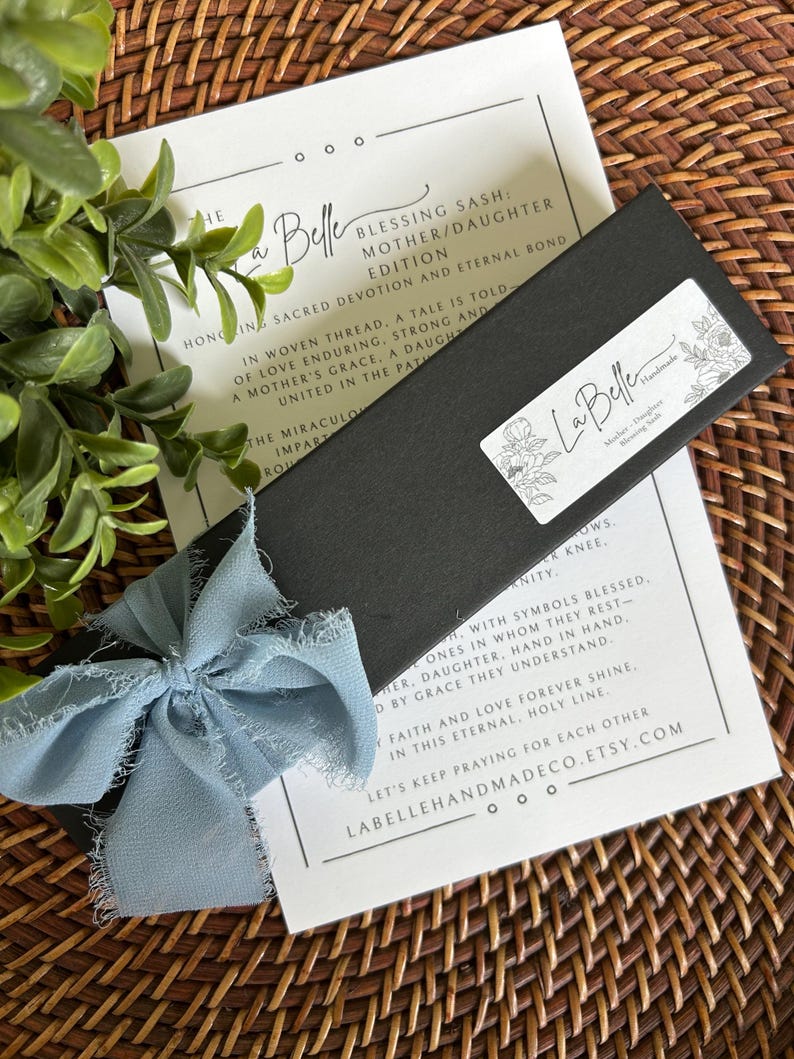 Puede incluir: Una caja negra con una cinta azul claro y una tarjeta con texto. La caja tiene una etiqueta blanca con dise&ntilde;os florales y las palabras "La Belle Handmade". La tarjeta dice "Blessing Sash: Mother/Daughter Edition".