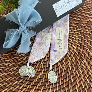 Puede incluir: Un marcap&aacute;ginas con estampado floral con dos medallas religiosas plateadas. El marcap&aacute;ginas es de color lavanda, blanco y dorado. Est&aacute; empaquetado en una caja negra atada con una cinta azul claro. La caja tiene una etiqueta que dice "La Belle Handmade".