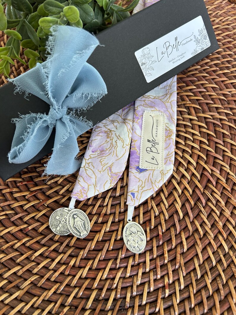 Puede incluir: Un marcap&aacute;ginas con estampado floral con dos medallas religiosas plateadas. El marcap&aacute;ginas es de color lavanda, blanco y dorado. Est&aacute; empaquetado en una caja negra atada con una cinta azul claro. La caja tiene una etiqueta que dice "La Belle Handmade".