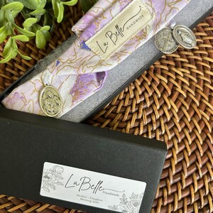 Puede incluir: Una faja con estampado floral en una caja negra. La faja es de color lila claro y blanco con detalles dorados. Dos colgantes plateados cuelgan de la faja. La caja tiene una etiqueta que dice "La Belle Handmade" y "Mother + Daughter Blessing Sash."